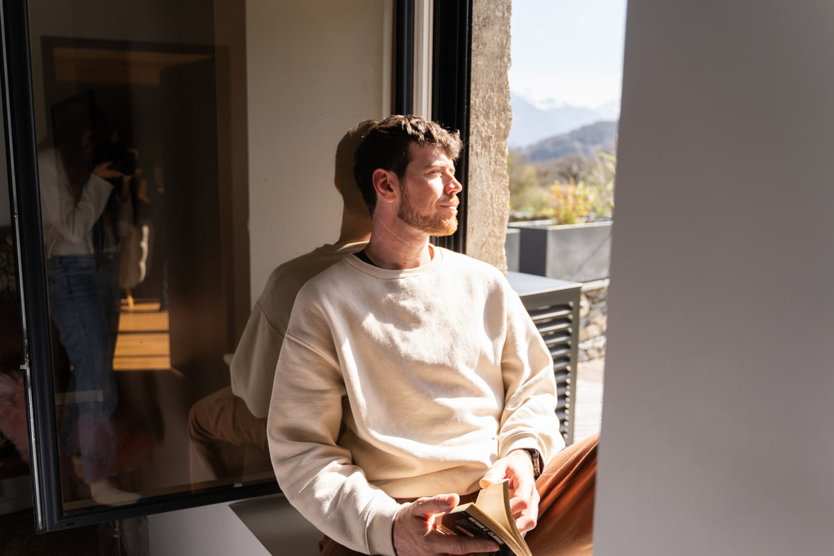 Ein Mann mit einem Buch in der Hand sitzt beim Fenster und blickt in die Natur raus. Ein Mann mit einem Buch in der Hand sitzt beim Fenster und blickt in die Natur raus.