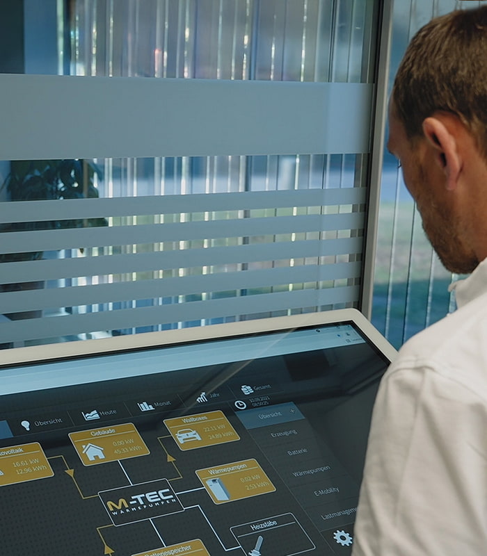 Ein Mann steht vor einem Touch Screen auf dem die Übersicht des Energiemanagements angezeigt wird. Ein Mann steht vor einem Touch Screen auf dem die Übersicht des Energiemanagements angezeigt wird.
