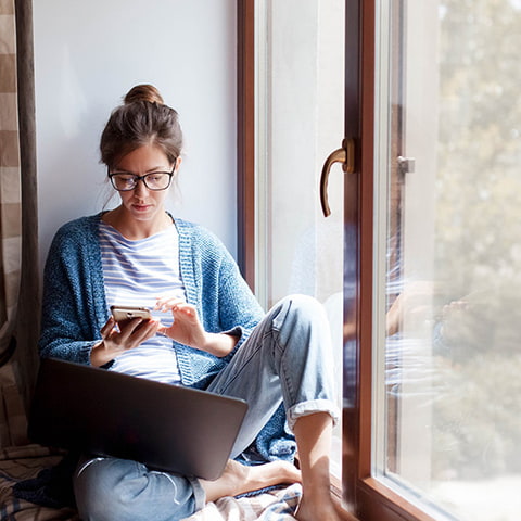 Ein Bild von einer jungen Frau mit Brille, die bei einem Fenster mit Handy und Laptop sitzt und sich informiert. Ein Bild von einer jungen Frau mit Brille, die bei einem Fenster mit Handy und Laptop sitzt und sich informiert.